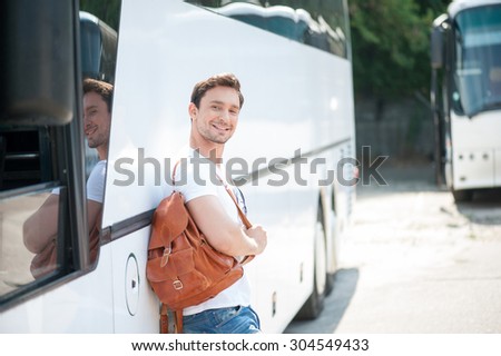 Handsome man is standing near a bus and leaning on it. He is holding a rucksack and smiling. The man is looking at the camera with joy. Copy space in right side