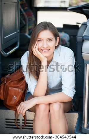 Pretty girl is sitting on the steps of the bus and waiting for its moving. She is looking at the camera and smiling. The lady is leaning her head on her hand
