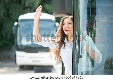 Cheerful young woman is standing on doorsteps of a bus. She is looking through the doors and smiling. She is waving her arm and greeting with people