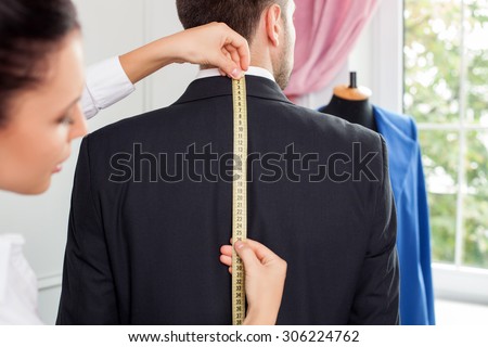 Beautiful female tailor is standing near her male client. She is measuring back of a man. The woman is looking at the tape-measure with concentration
