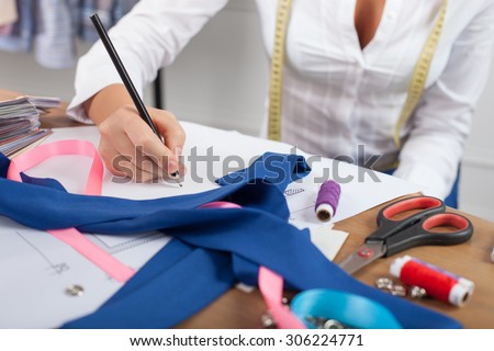Close up of hands of skilled fashion designer. The woman is sitting at the table. She is drawing sketches of clothes on a blueprint. There are many design things on the desk
