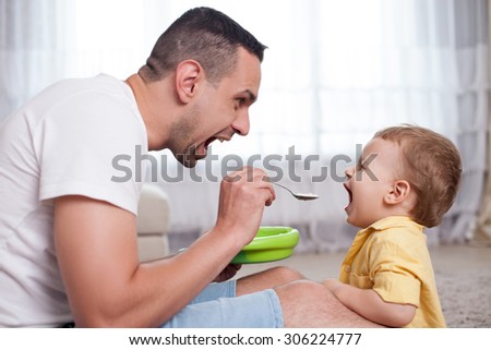 Cheerful father is teaching his little son to eat porridge from the spoon. They are opening their mouths widely. The family is sitting on flooring with joy