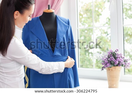 Beautiful clothes designer is sewing a belt for modern jacket. She is measuring it with tape-line. The woman is standing near a mannequin and smiling