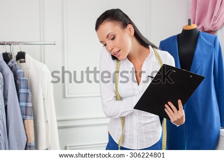 Cheerful clothes designer is talking on the phone with her client. She is holding a folder of papers and writing down some data. The woman is smiling. She has a tape measure over her neck