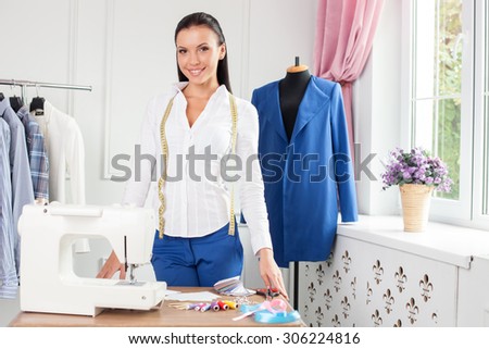 Beautiful clothes designer is creating new clothing. She is standing near a sewing machine and mannequin. The woman is leaning on the table and smiling. She has a tape-line over her neck