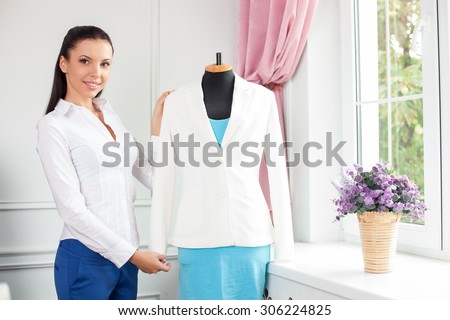 Attractive clothes designer is presenting her new clothing. She is standing near a mannequin and smiling. The woman is looking forward with happiness