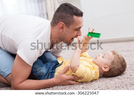 Attractive man is playing with his son on flooring. The boy is holding a toy and lying on carpet with joy. His parent is looking at him playfully and smiling