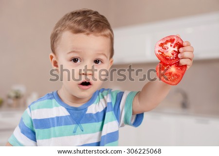 Cheerful small boy is holding pieces of chopped tomato. He is showing it to the camera and looking forward with interest