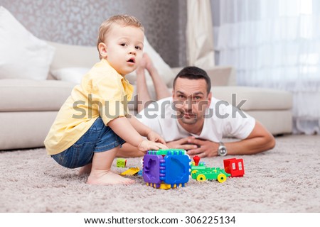Cheerful father is lying on flooring near his son and smiling. They are playing with toys. They boy is sitting on a carpet and looking at the camera with interest
