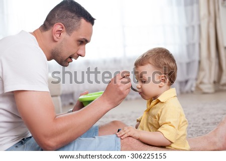 Handsome father is feeding his child with porridge. He is looking at the kid with love. The boy is eating food with appetite. The family is sitting on flooring