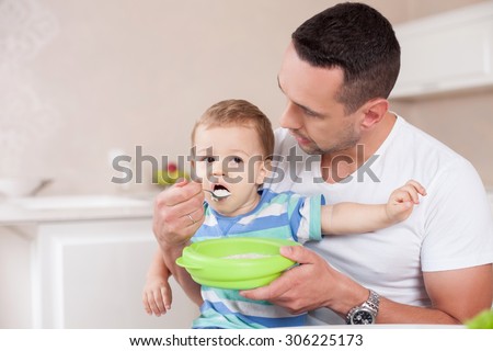 Handsome father is feeding his son with concentration. He is sitting in the kitchen and holding the boy on his knees. The toddler is eating porridge with appetite. Copy space in left side