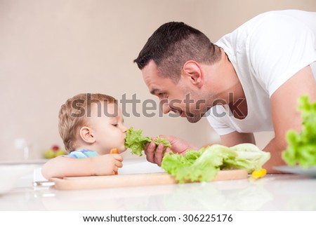 Cute young man is feeding his son with lettuce. He is looking at the toddler and smiling. The boy is eating vegetable with appetite. He is sitting at the table and holding a carrot