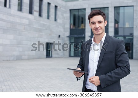 Handsome man in suit is standing near his office and smiling. He is holding a tablet and looking at the camera with joy. Copy space in left side