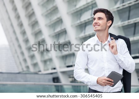 Cheerful businessman is standing and leaning on the border of building. He is holding tablet and his jacket behind his back. The worker is smiling and looking aside dreamingly. Copy space in left side