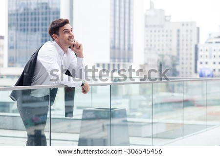 Cheerful businessman is talking on the mobile phone outdoors. He is standing and leaning on the border. The worker is holding his jacket behind his back and smiling. Copy space in right side