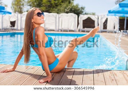 Beautiful girl is sitting near a swimming pool. She is putting her legs in pool and splashing water. The lady is smiling. She closed her eyes with pleasure