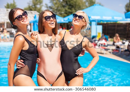 Cheerful women are standing near a swimming pool and relaxing. They are embracing and looking at the camera with happiness. The friends are posing and smiling. Copy space in right side