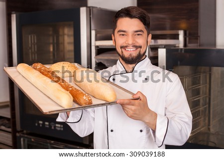 Handsome chef is standing in bakery and smiling. He is holding a tray with baked products and showing it to camera proudly