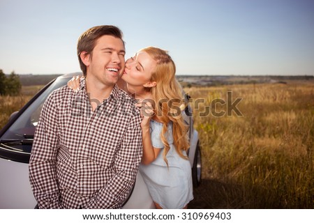 Cheerful loving couple is standing near their transport in the nature. The woman is embracing and kissing her boyfriend with love. The man is smiling with happiness. Copy space in right side
