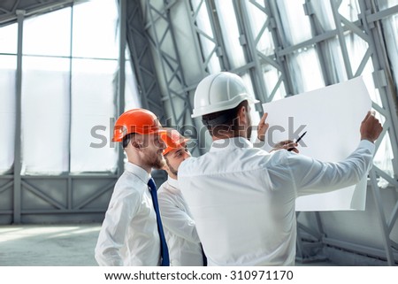 Pretty architect is explaining to builders the plan of construction. He is holding a pen and pointing it at the blueprint with seriousness. The foreman is smiling. Copy space in left side