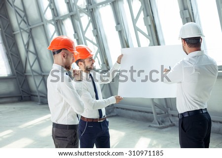 Handsome builders are holding a blueprint and looking at it seriously. They are discussing the plan of a new building. The engineer is pointing at paper with explanations. His colleague is smiling