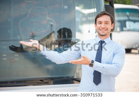 Attractive driver is standing near a bus and presenting it. He is raising his hands sideways and smiling