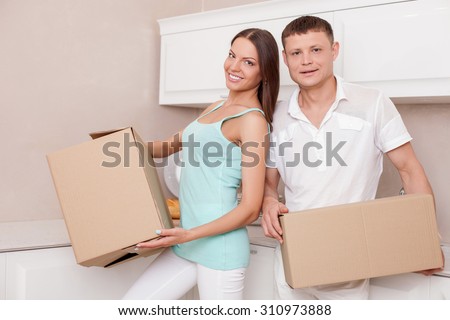 Cute young married couple is standing in kitchen and holding boxes in their hands. They are moving in a new building. They are smiling happily and looking at the camera with joy