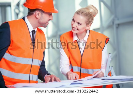 Cheerful architects are standing near a table with blueprint on it. The woman is pointing her finger at sketches. She is explaining her ideas about building to her colleague. They are smiling