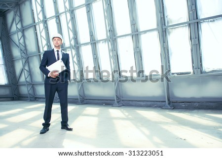 Successful engineer is standing in large empty building. He is waiting for his customer. The man is holding a folder of documents and looking forward with seriousness. Copy space in right side