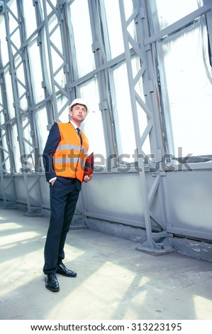Handsome architect is standing in workwear a white helmet. He is looking forward seriously. The man is holding a folder and putting arm in his pocket. Copy space in right side