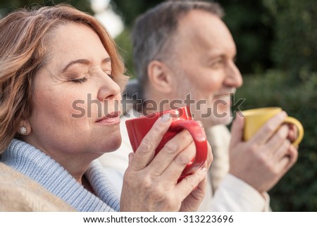 Cheerful old married couple is drinking hot tea. They are sitting on grass and smiling. The woman closed her eyes with relaxation
