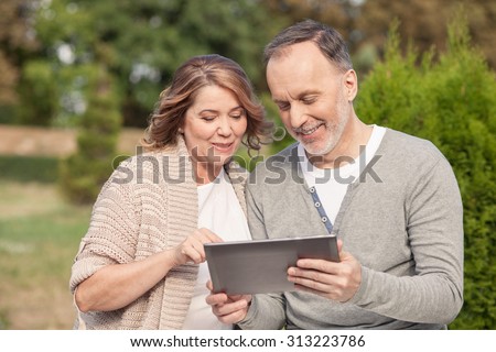 Cute mature married couple is looking at the laptop happily. The man is holding it in his hands. The woman is touching the technology. They are smiling