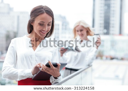 Attractive young woman is standing on the balcony of her office. She is using a tablet and smiling. Her female colleague is standing behind her and reading newspaper with interest
