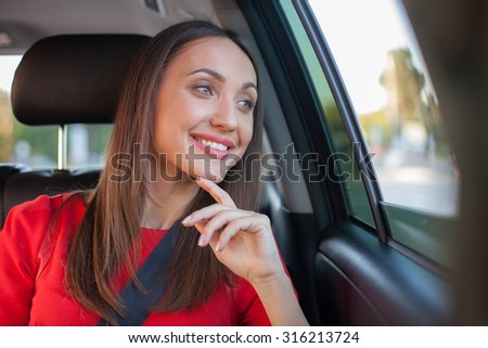 Beautiful businesswoman is sitting on back seat of the car. She is looking through the window dreamingly and smiling. The lady is touching her chin with excitement