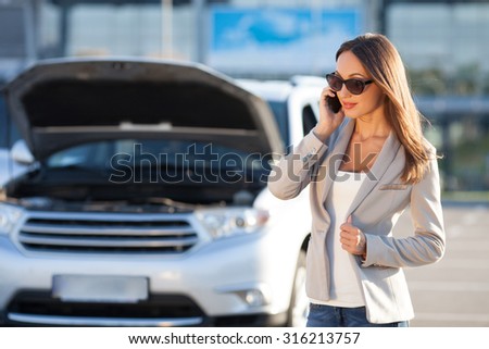Attractive young woman is standing near her broken car. She needs for help. The girl is talking on the mobile phone and smiling. The bonnet of her car is open