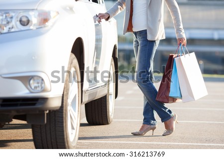 Close up of body of woman standing near her car. She is touching the handle and opening the door of vehicle. The lady is holding packets after shopping