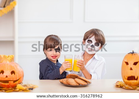 Cute boy and girl are making fun in Halloween. They are sitting in kitchen and drinking juice. The friends have spooky make-up on their faces and smiling. There are pumpkins and cookies on the table