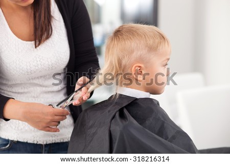 Skilled female barber is making a haircut for boy with concentration. The kid is sitting in chair with seriousness. The woman is holding a comb and scissors