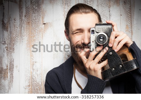 Attractive bearded man is photographing with interest. He is holding camera near his eye and smiling. The guy is standing near wall. Copy space in left side