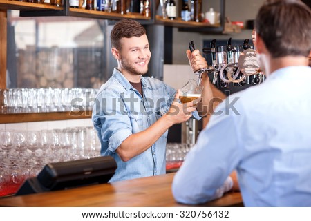 Cheerful male barman is serving his customer. He is pouring beer in glass and smiling. The man is sitting at counter and waiting for his order