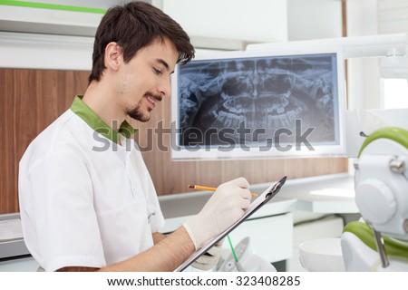 Cheerful young dental doctor is working in laboratory. He is holding a folder of document and writing. The man is sitting near x-ray of human jaw on screen. He is smiling