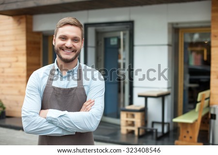 Handsome young barista is standing near his cafe outdoors. He is looking forward and smiling. The man crossed his arms with confidence