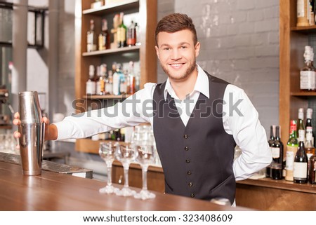 Attractive male bartender is holding a shaker and mixing drink. He is standing at counter near glasses. The man is looking forward and smiling