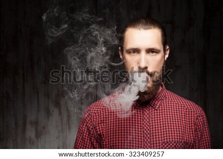 Handsome bearded man is standing and breathing out smoke. He is looking forward seriously. Copy space in left side