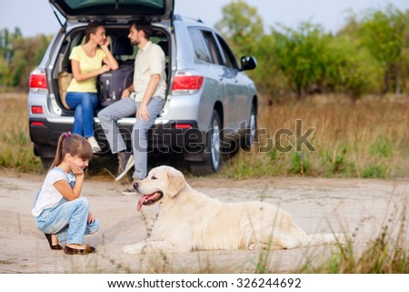Cheerful small girl is playing with dog in the nature. Her parents are sitting in open car boot and talking. The man and woman are looking at each other with love. They are smiling