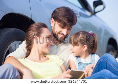 Beautiful young married couple with small daughter are sitting near car. They are making trip with joy. The father is embracing his wife and girl. They are reading book and smiling