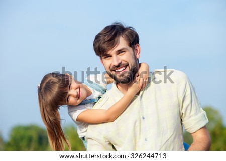 Attractive young man and his daughter are resting in the nature. The father is standing and carrying kid on his back. The girl is embracing him. They are looking forward and smiling