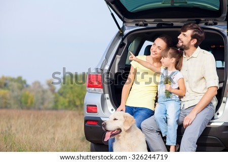 Cheerful young married couple and daughter are sitting on car trunk near dog. They are enjoying nature and smiling. The girl is pointing finger sideways. Parents are looking there with interest