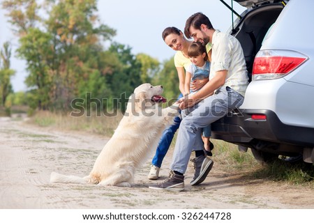 Pretty family is resting in forest and playing with dog. They are sitting on open car boot and embracing. The man and woman with girl are smiling. Copy space in left side