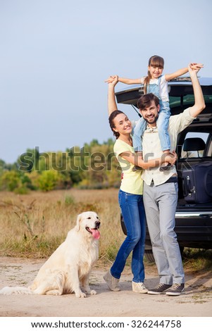 Cute family is resting in the nature and smiling. They are standing near car and dog. The father is holding daughter on shoulders. His wife is embracing him with love